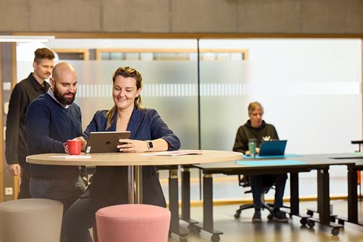 Links im Bild sind zwei Personen im Vordergrund an einem Stehtisch und unterhalten sich über ein Tablet gebeugt. Hinter ihnen geht ein junger Mann entlang. Rechts im Bild ist im Hintergrund ein älterer Mann an einem Schreibtisch zu sehen.